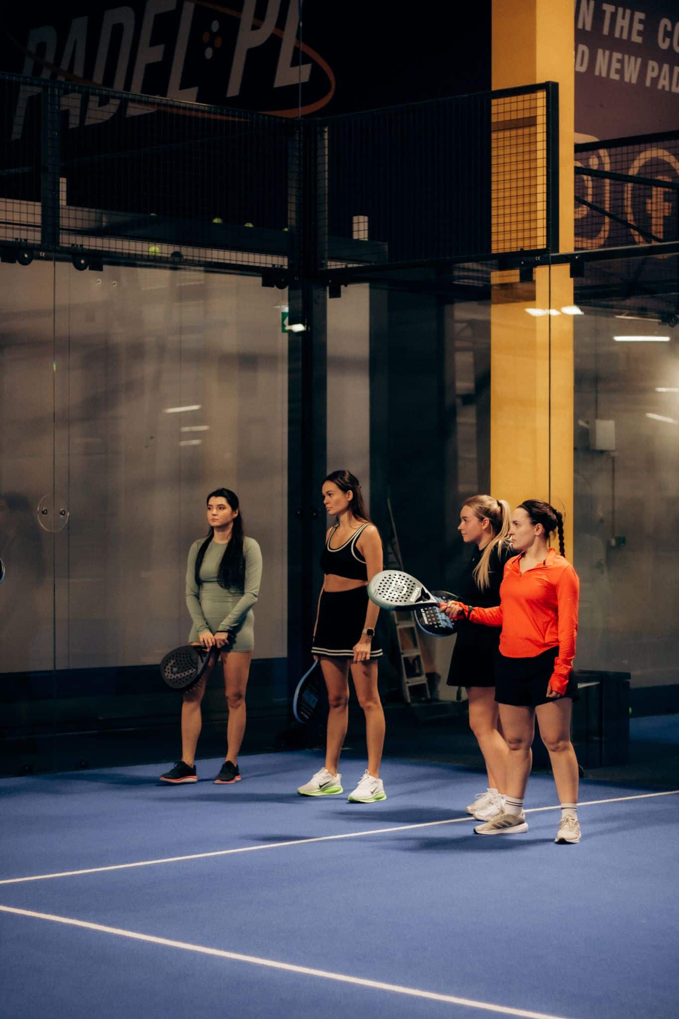 Four women holding rackets stand on a blue padel court, ready to play indoors.