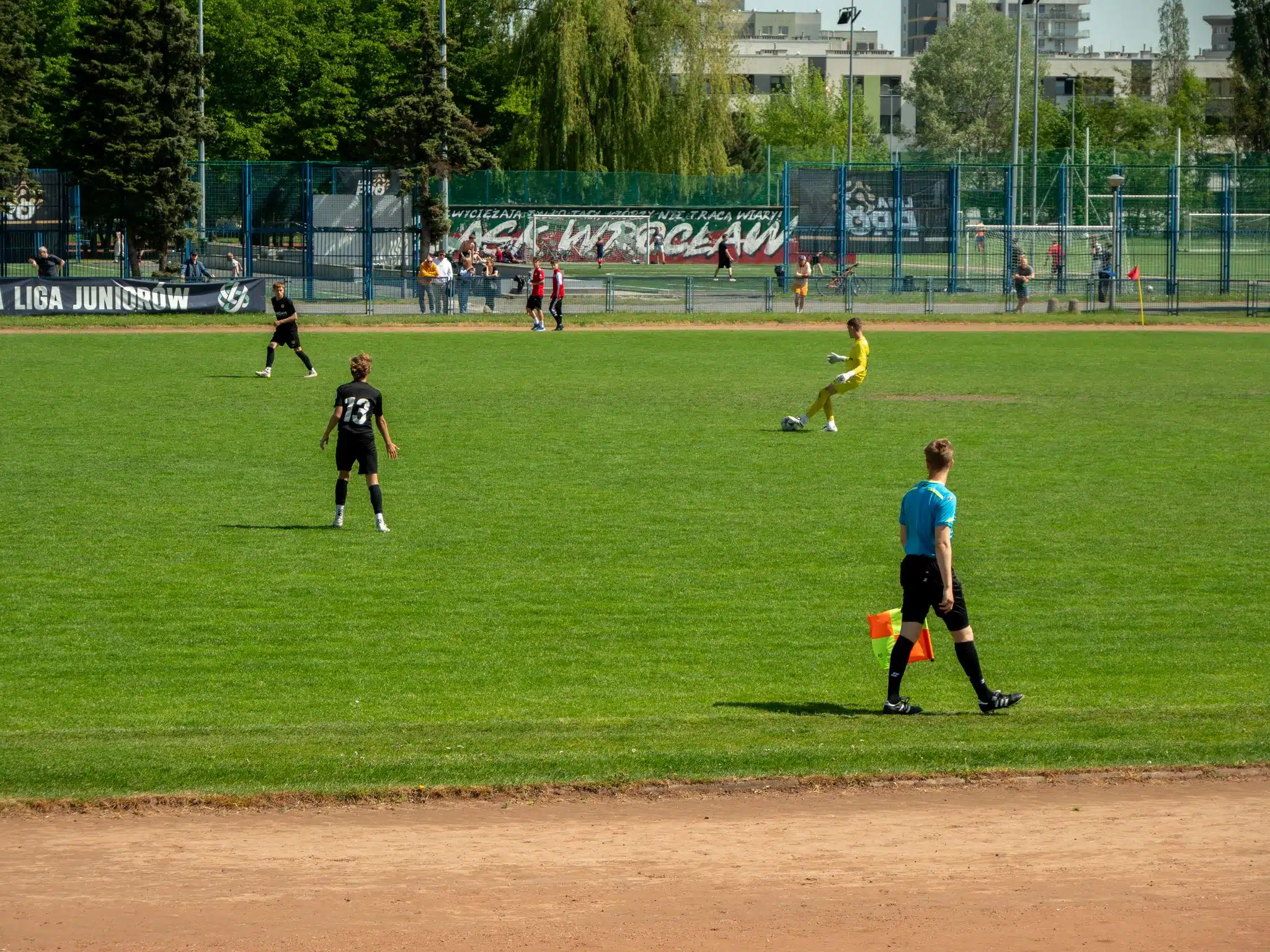 Youth soccer game at Wrocław sports field on a sunny day with players and referee in action.